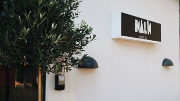 A sign for 'KALM HOTEL' mounted on a white wall, accompanied by a potted plant and a wall-mounted light fixture.