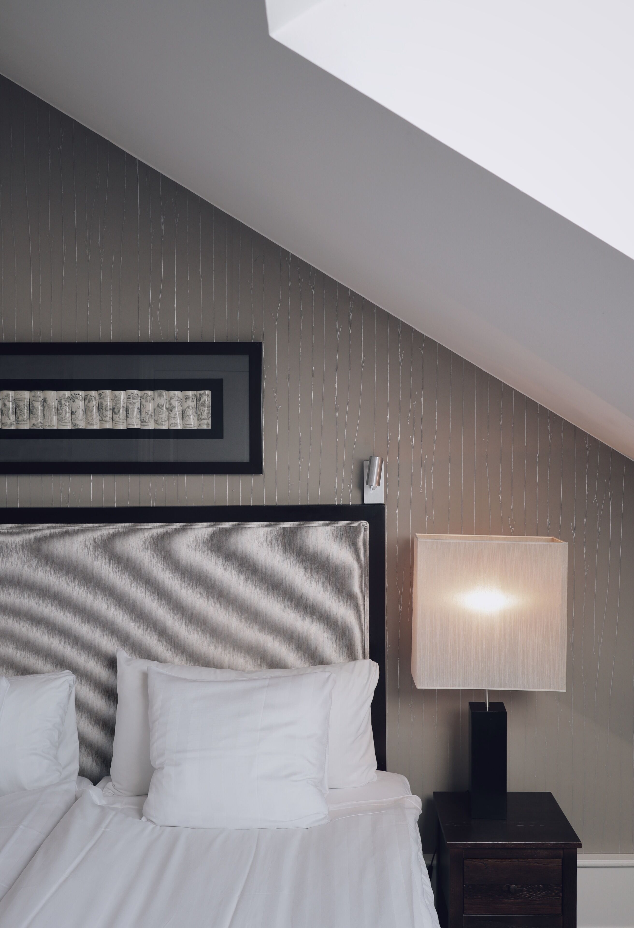 A hotel room with a bed, a nightstand with a lamp, a framed picture on the wall, and a sloped ceiling.