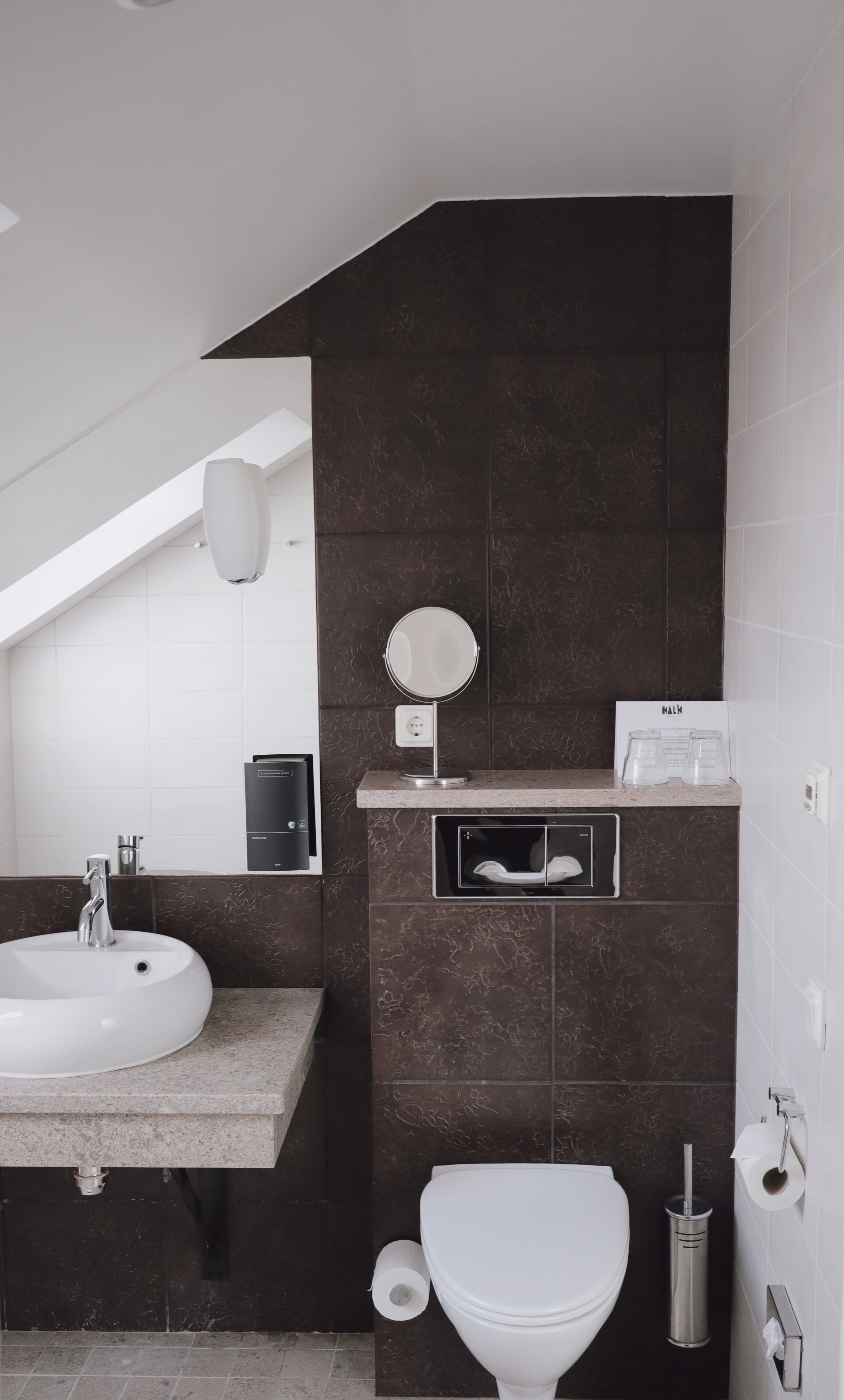 Une salle de bain moderne avec un lavabo blanc, des murs recouverts de carreaux foncés, des toilettes et un petit miroir.