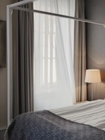 A bedroom with a four-poster bed, sheer curtains, a bedside lamp, and a view of the outside.