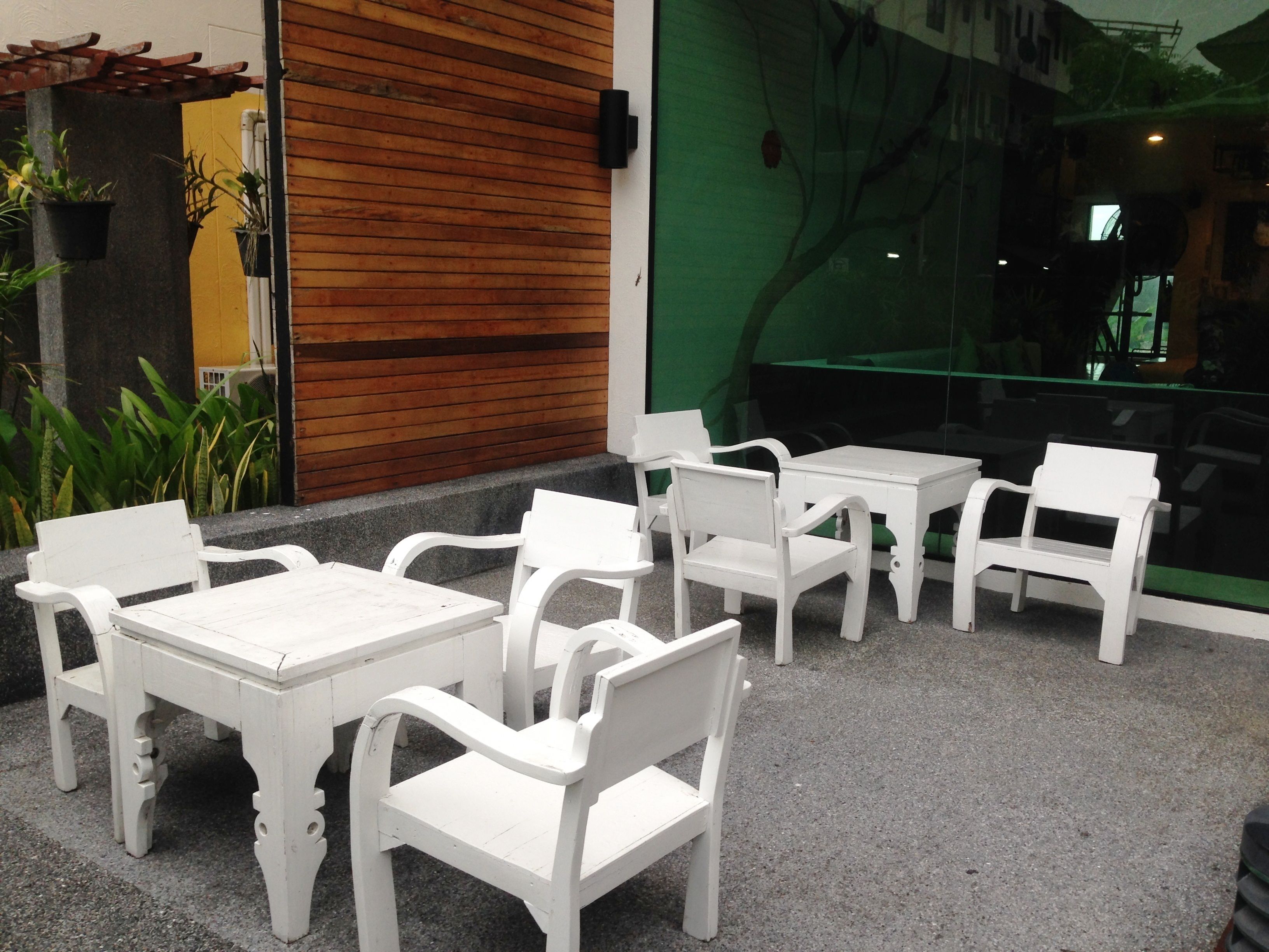 Outdoor seating with white tables and chairs, a wooden wall, and a glass window with a green view.