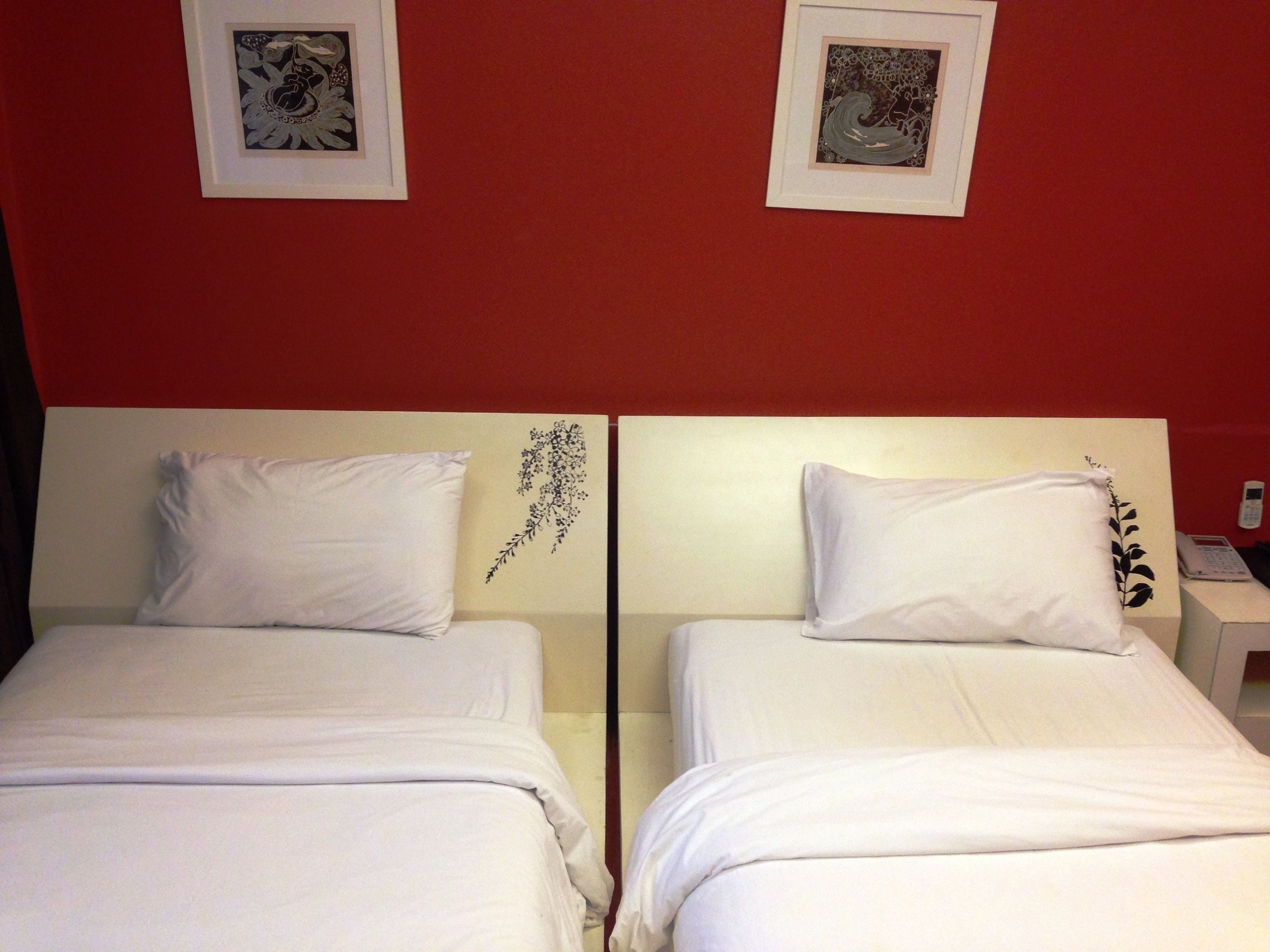 Two single beds with white linens, a red wall, and two framed artworks above the beds.