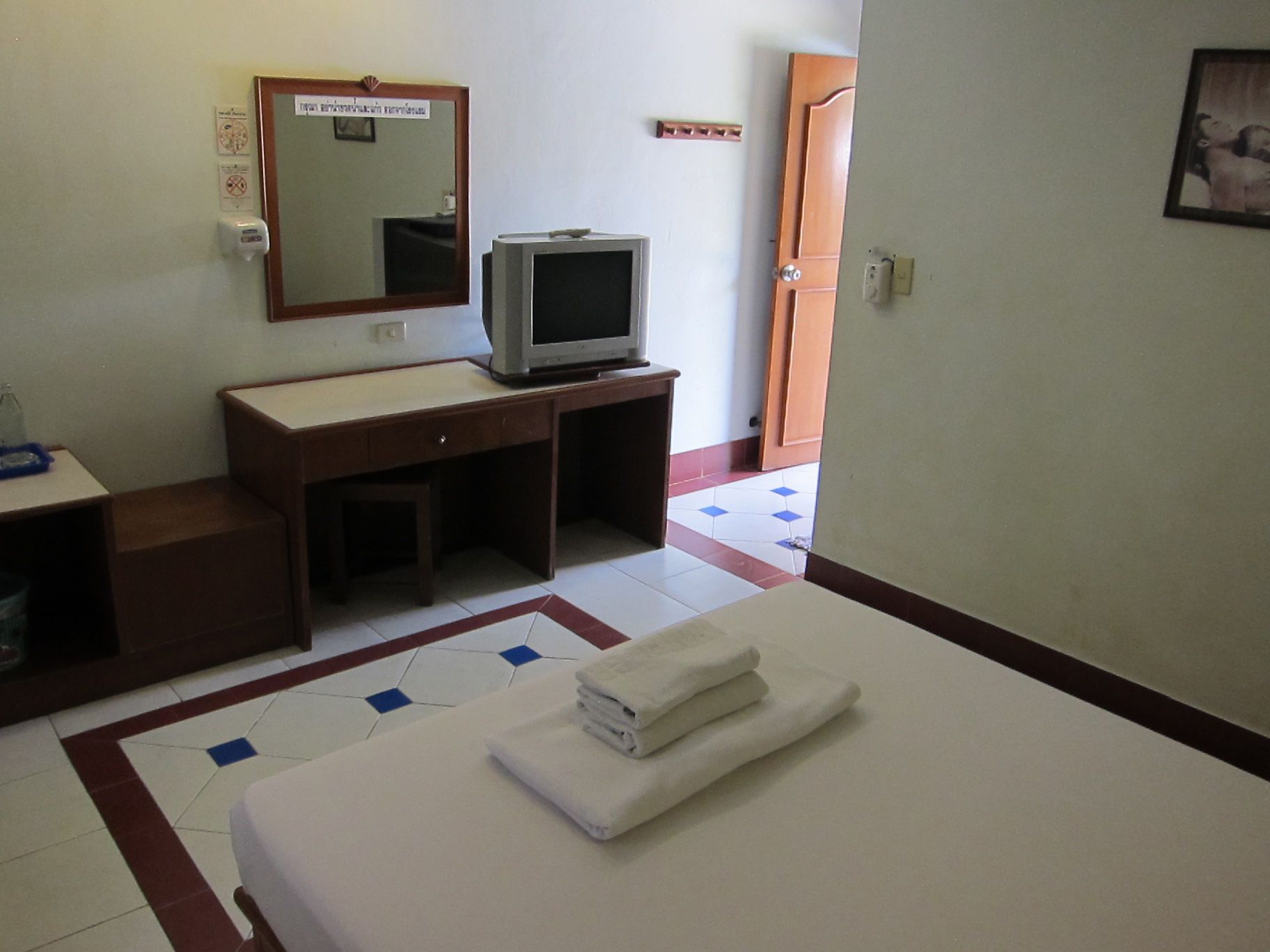Una camera d'albergo con un letto, una TV su un supporto in legno, uno specchio e un pavimento piastrellato.