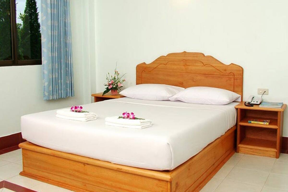 A neatly made bed with white linens and two towels, a wooden headboard, a bedside table with a phone and a book, and a window with a curtain.