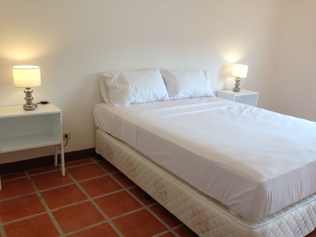 Habitación con cama individual, ropa de cama blanca, dos mesitas de noche con lámparas y suelo de baldosas.