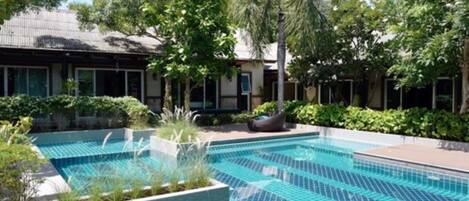 Una piscina con bordes de azulejos, rodeada de vegetación y un edificio con grandes ventanales.