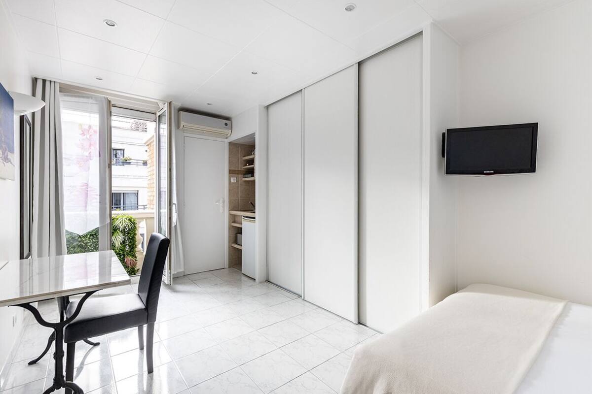 Una habitación de hotel moderna con cama, televisor montado en la pared, mesa de comedor con sillas y puerta corrediza que da a un balcón.