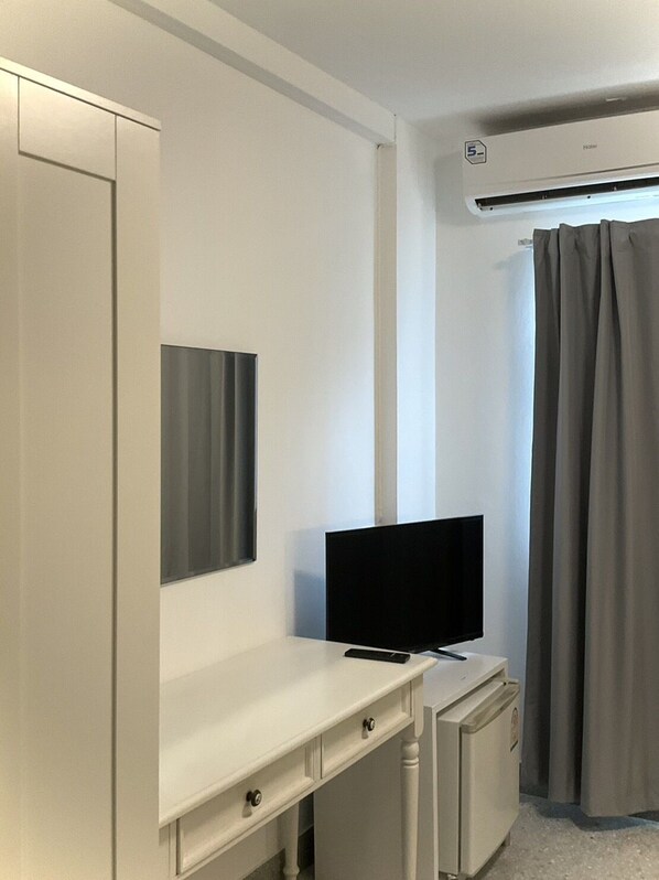 A room with a white desk, a flat-screen TV, a small refrigerator, and a window with grey curtains.