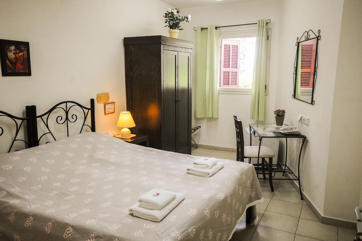Un lit bien fait, recouvert d’une couvre-lit à motifs floraux, avec deux serviettes pliées, une lampe de chevet, une armoire en bois foncé, une petite table avec un téléphone et un pot de fleurs, et une fenêtre ornée de rideaux verts.