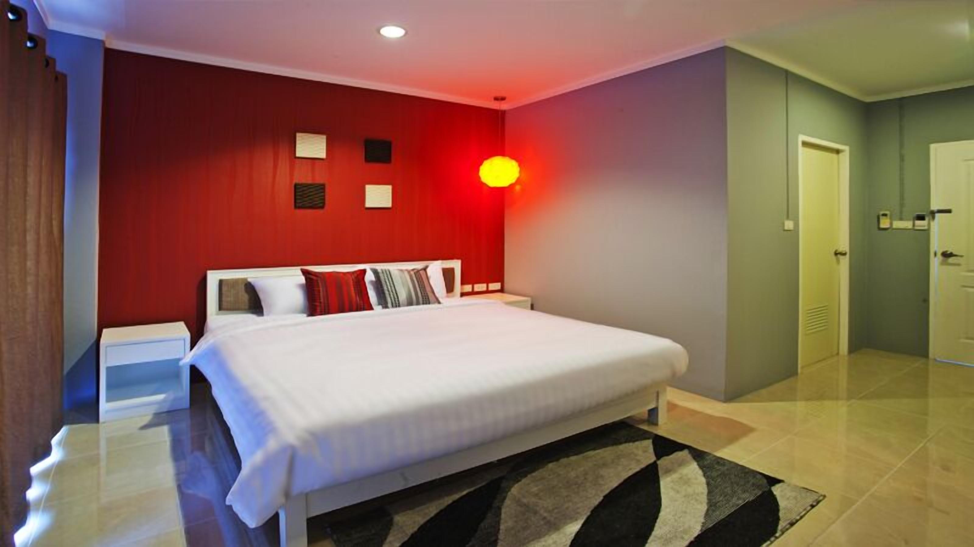 Una habitación de hotel con una cama grande, una mesita de noche, una pared de acento roja, una lámpara fijada a la pared y una puerta.