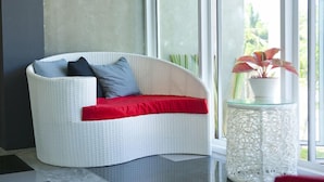 A white wicker sofa with red and grey cushions, positioned next to a glass table with a white, textured base, near a large window with a view of greenery. - I Style Hua Hin Hotel (Hua Hin)