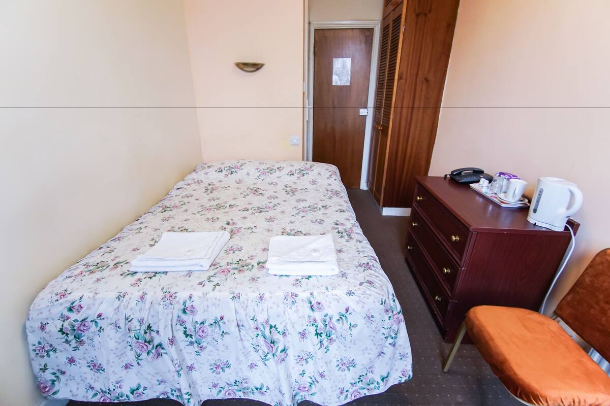 A neatly made bed with a floral bedspread, a wooden dresser with a kettle, and a chair.