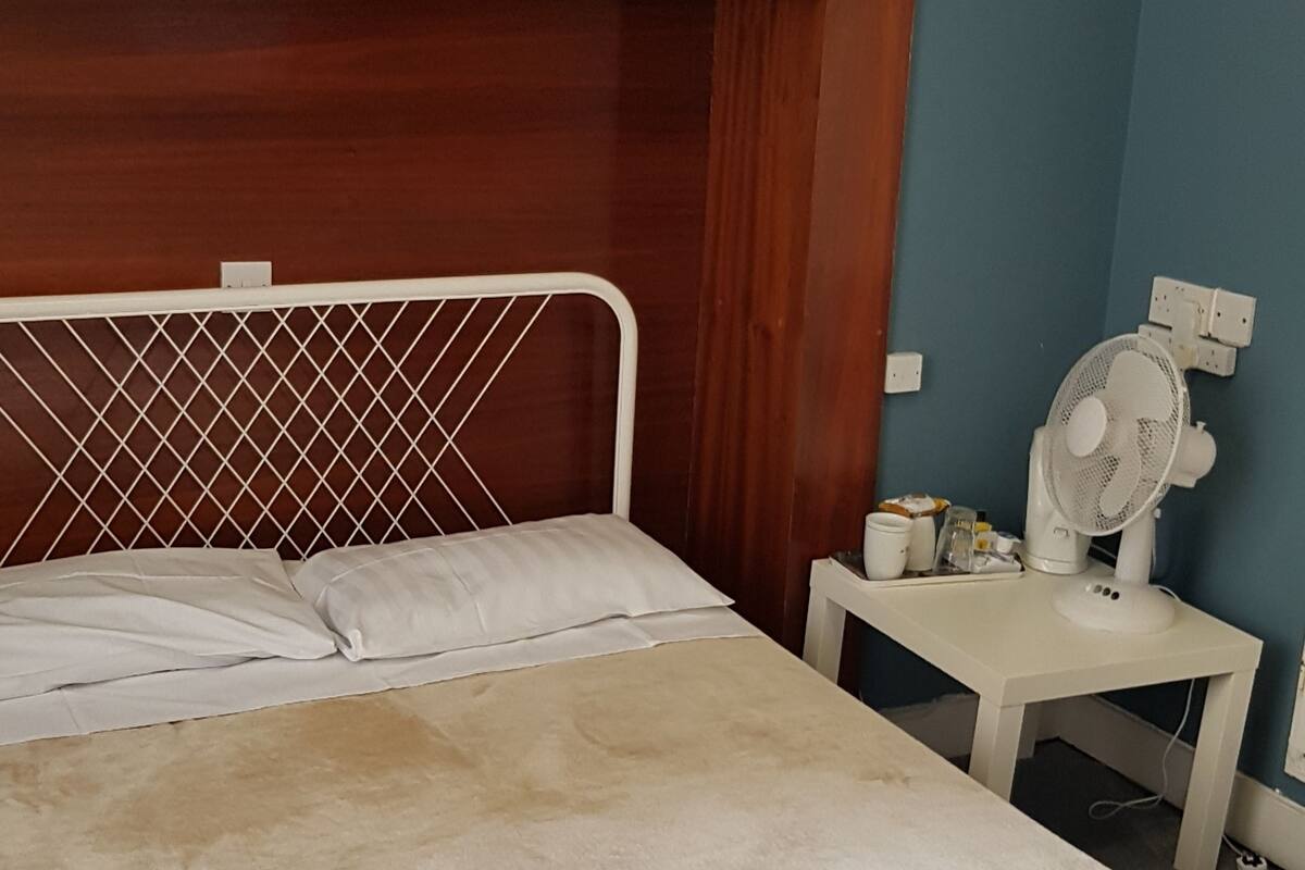 A bedroom with a wooden headboard, a white mattress, a small bedside table with a fan, and a wall socket.