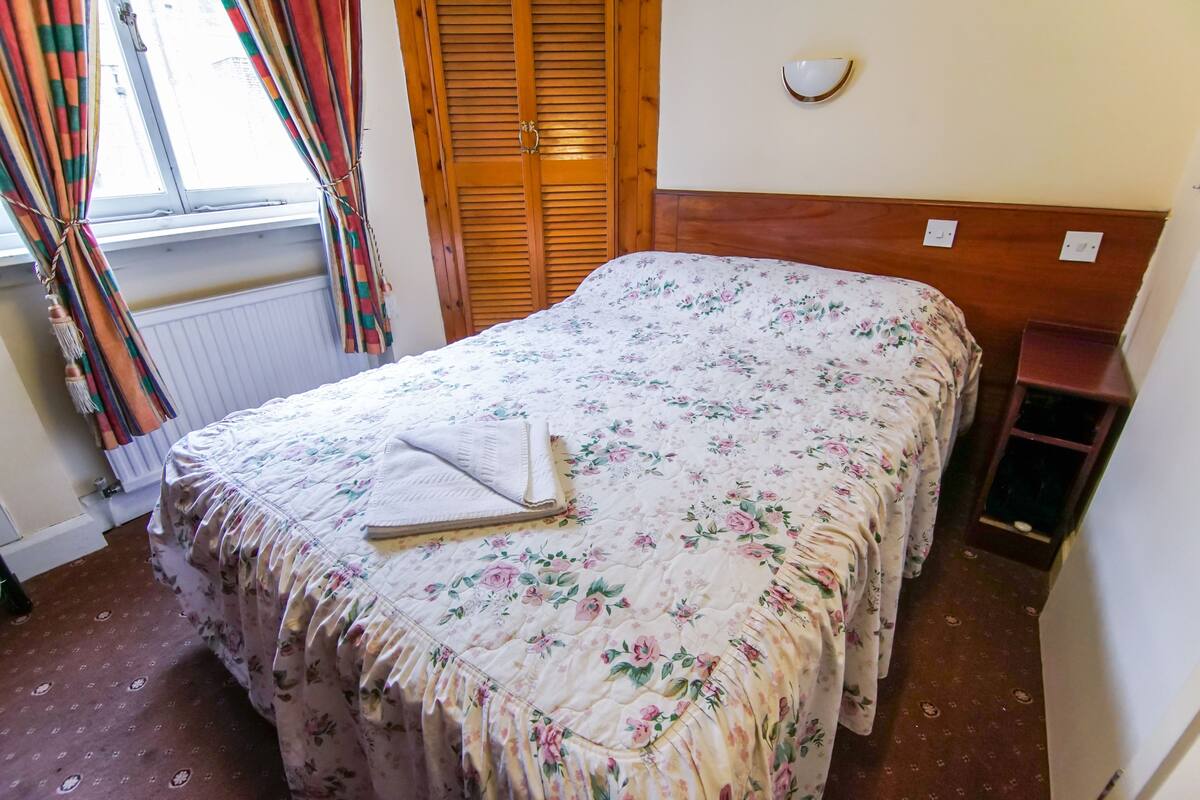 A neatly made bed with a floral bedspread, a wooden headboard, a bedside table, and a window with curtains.