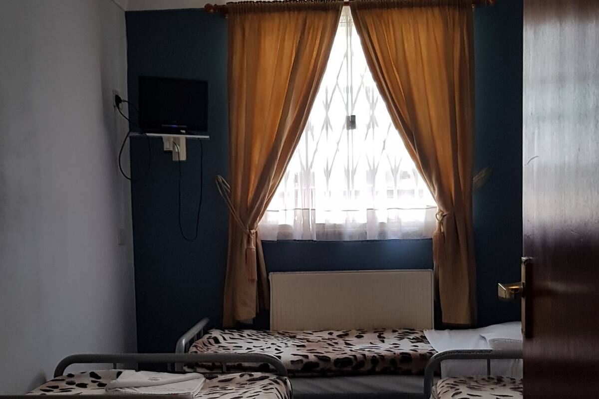 A small, dimly lit room with two single beds, a television on the wall, and a window with curtains.