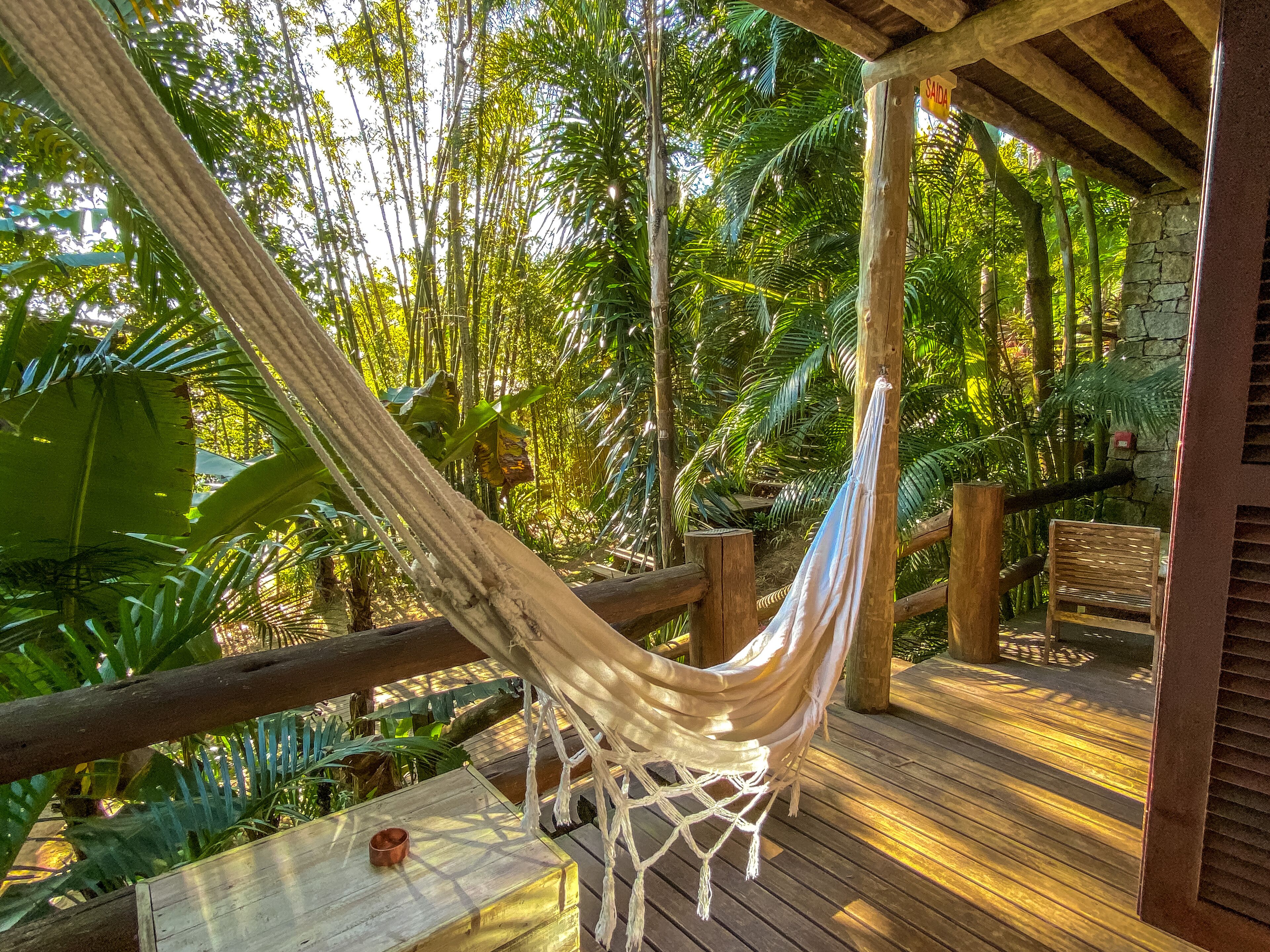 A hammock hanging between wooden beams in a lush, tropical garden with a wooden deck and stone walls.