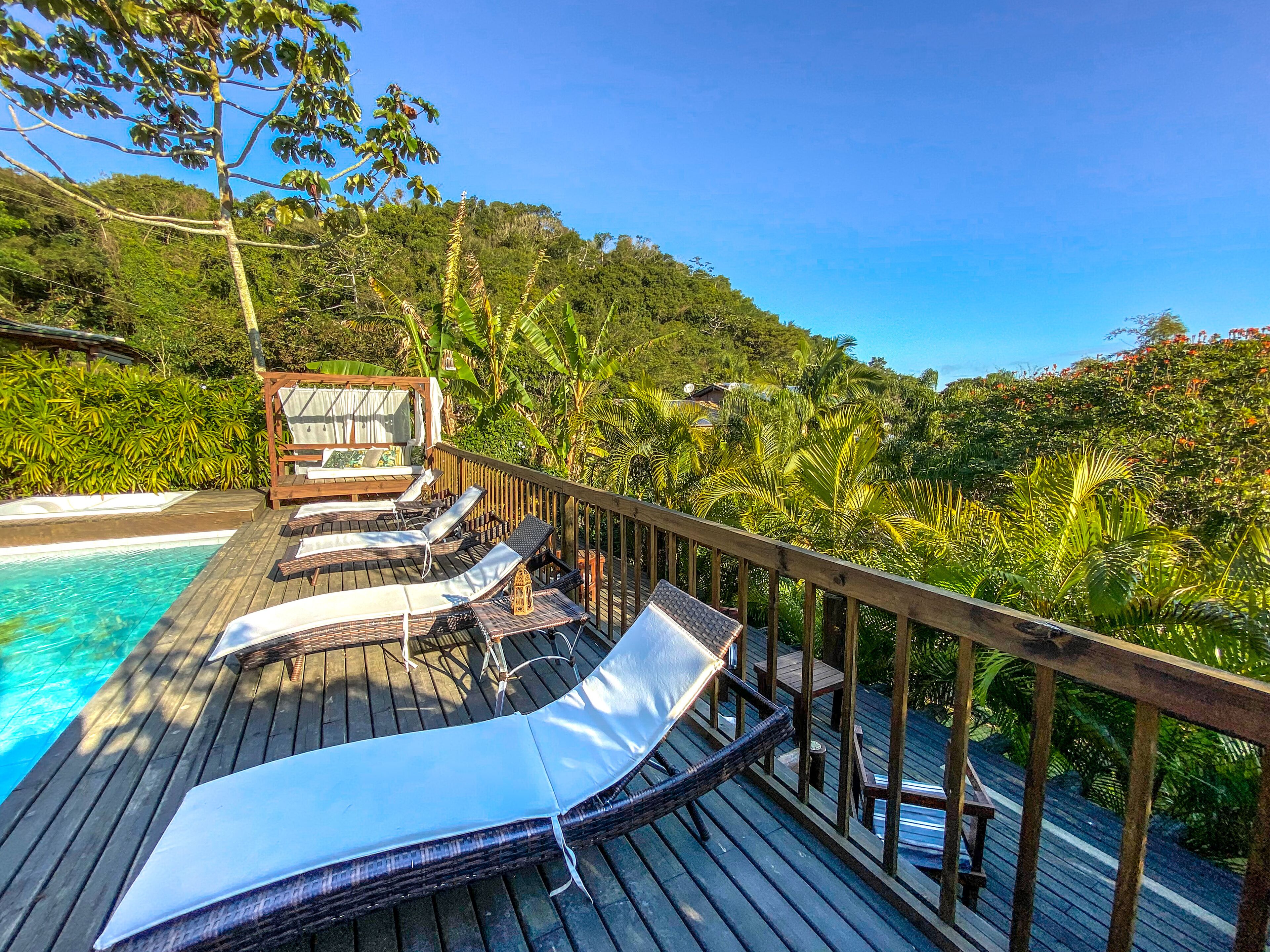 A wooden deck with lounge chairs and a hot tub overlooking a pool and lush greenery.