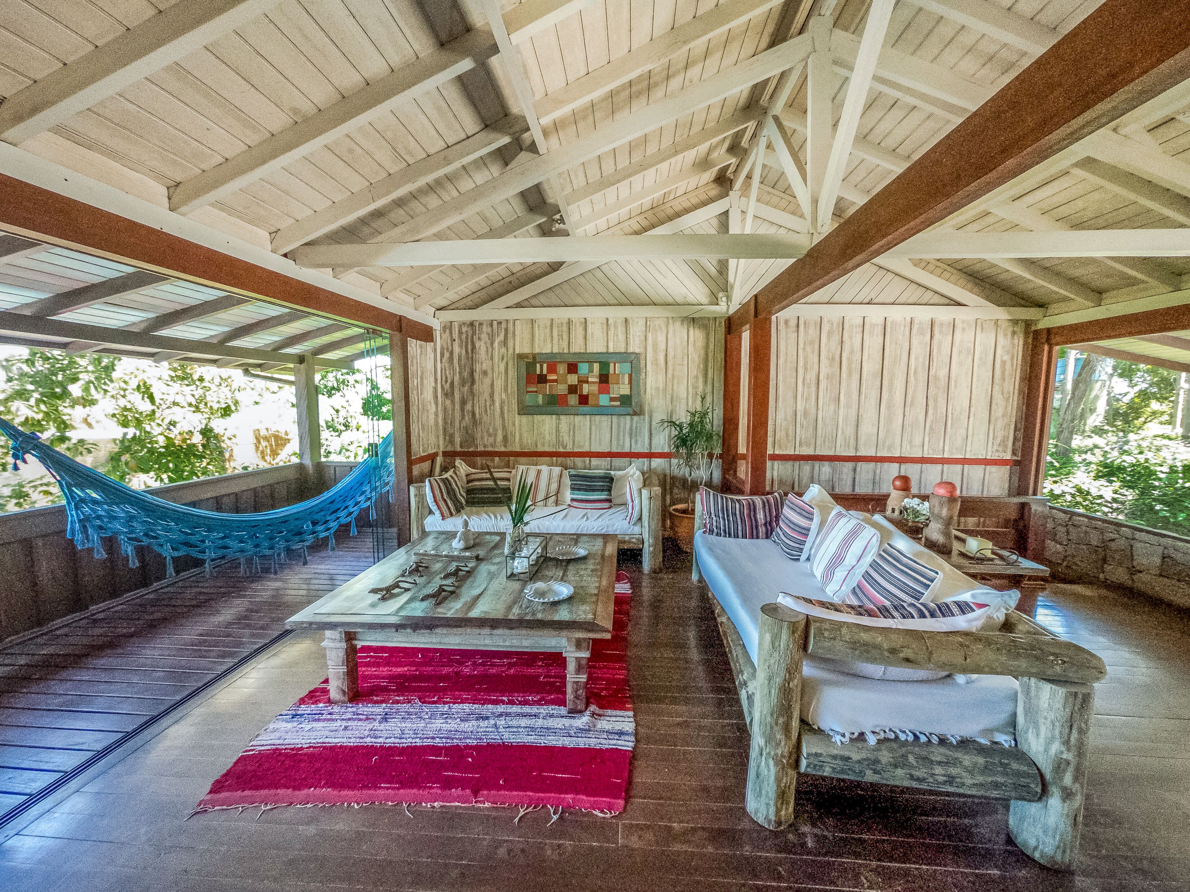 A spacious room with a high ceiling, wooden beams, a hammock, a sofa, a coffee table, and a red rug.