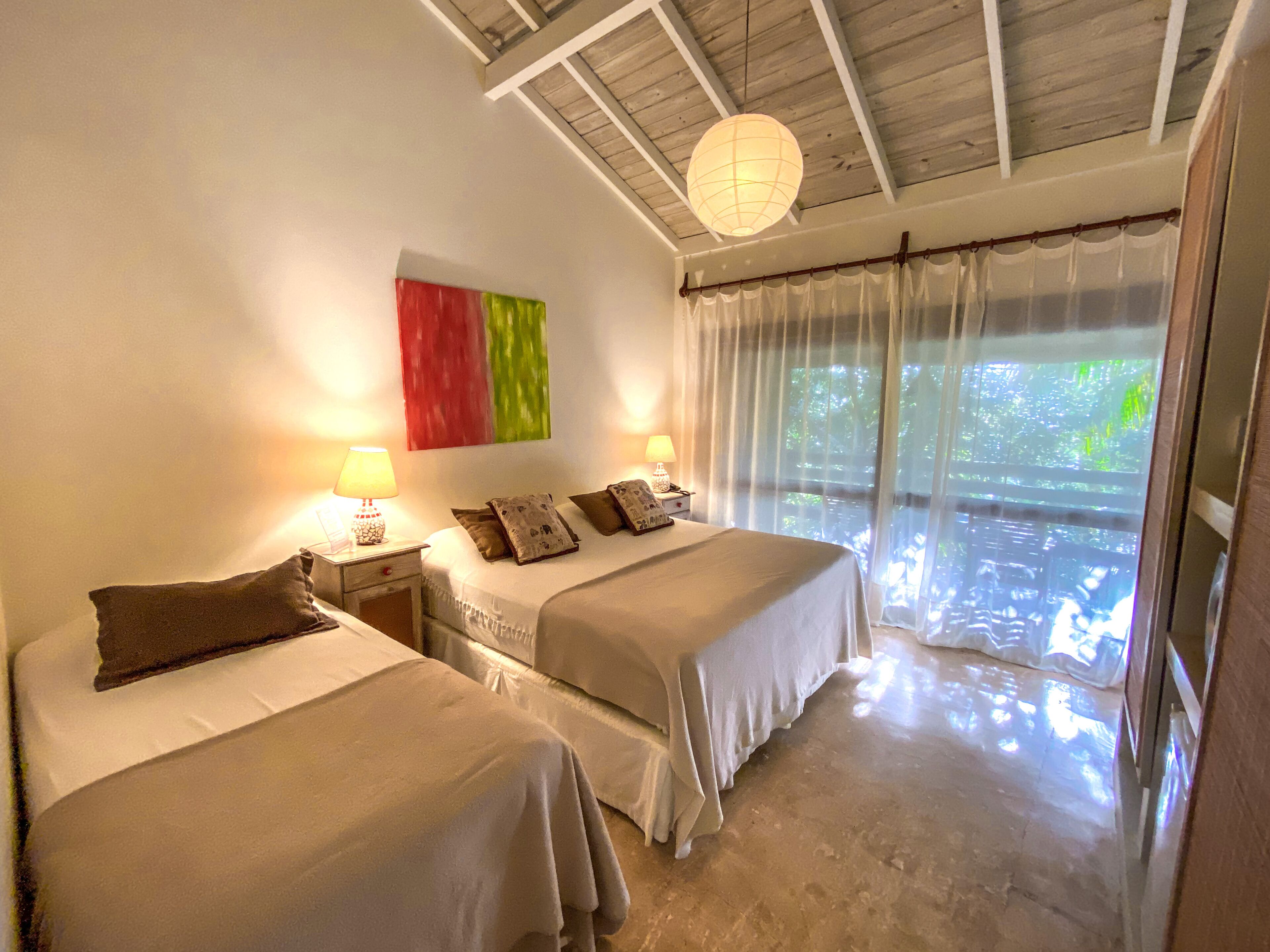 A bedroom with two beds, a nightstand with a lamp, a red and green artwork on the wall, and a view of greenery through sheer curtains.