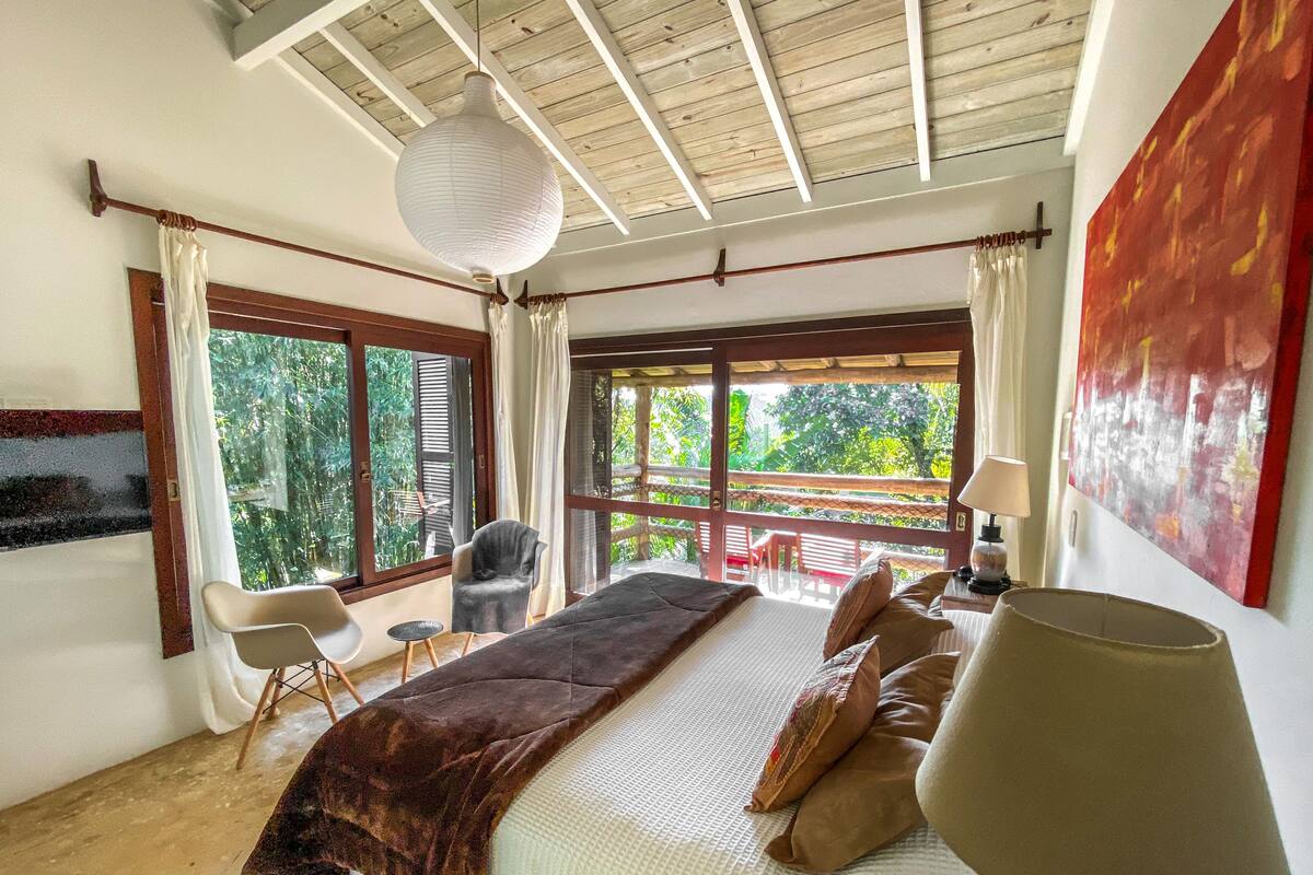 A bedroom with a large bed, a hanging light fixture, a view of the outdoors, and a painting on the wall.