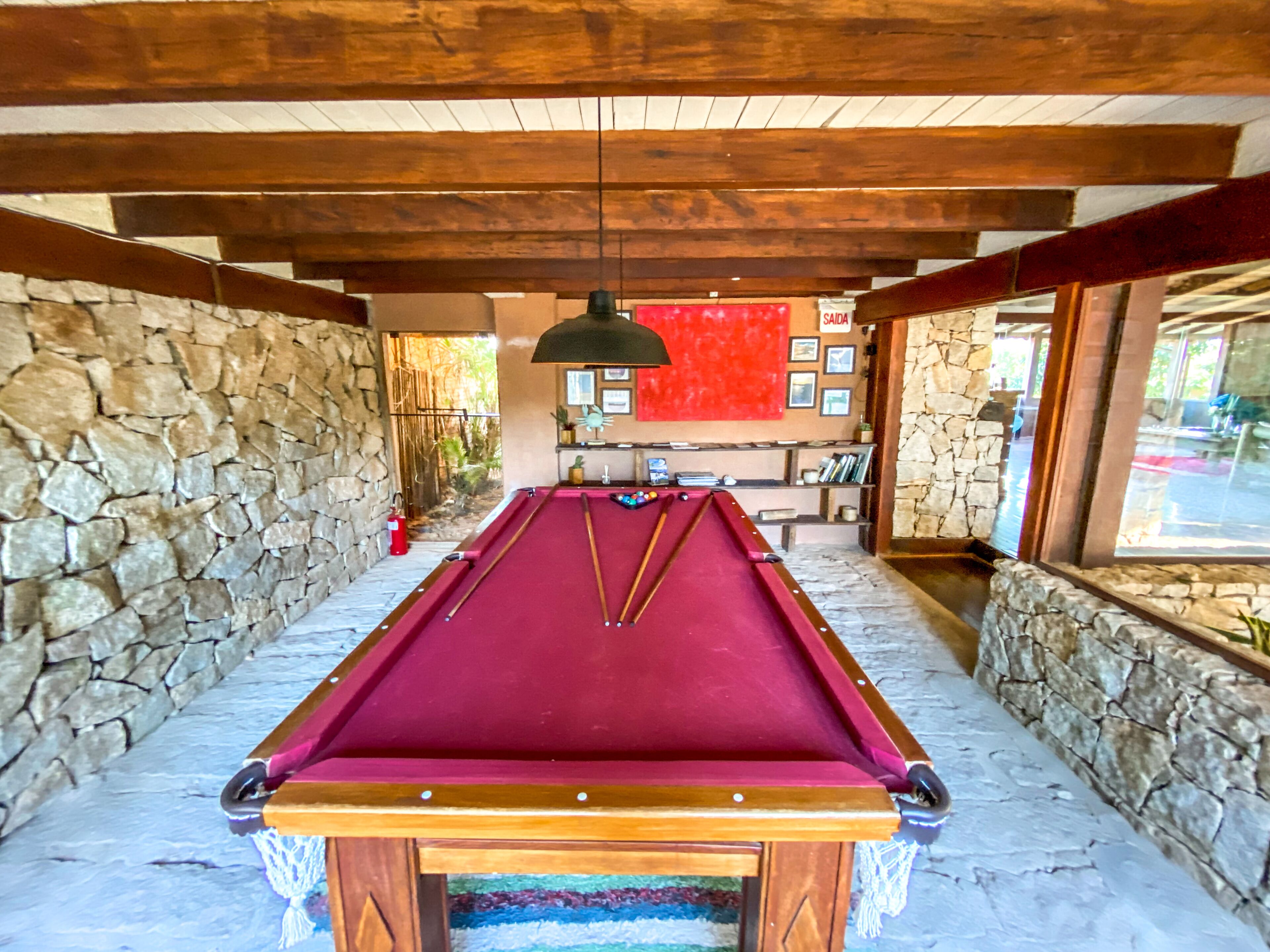 A pool table in a room with stone walls and wooden beams, a red wall with framed pictures, and a window with a view.