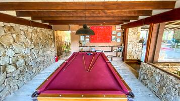 A pool table in a room with stone walls and wooden beams, a red wall with framed pictures, and a window with a view.