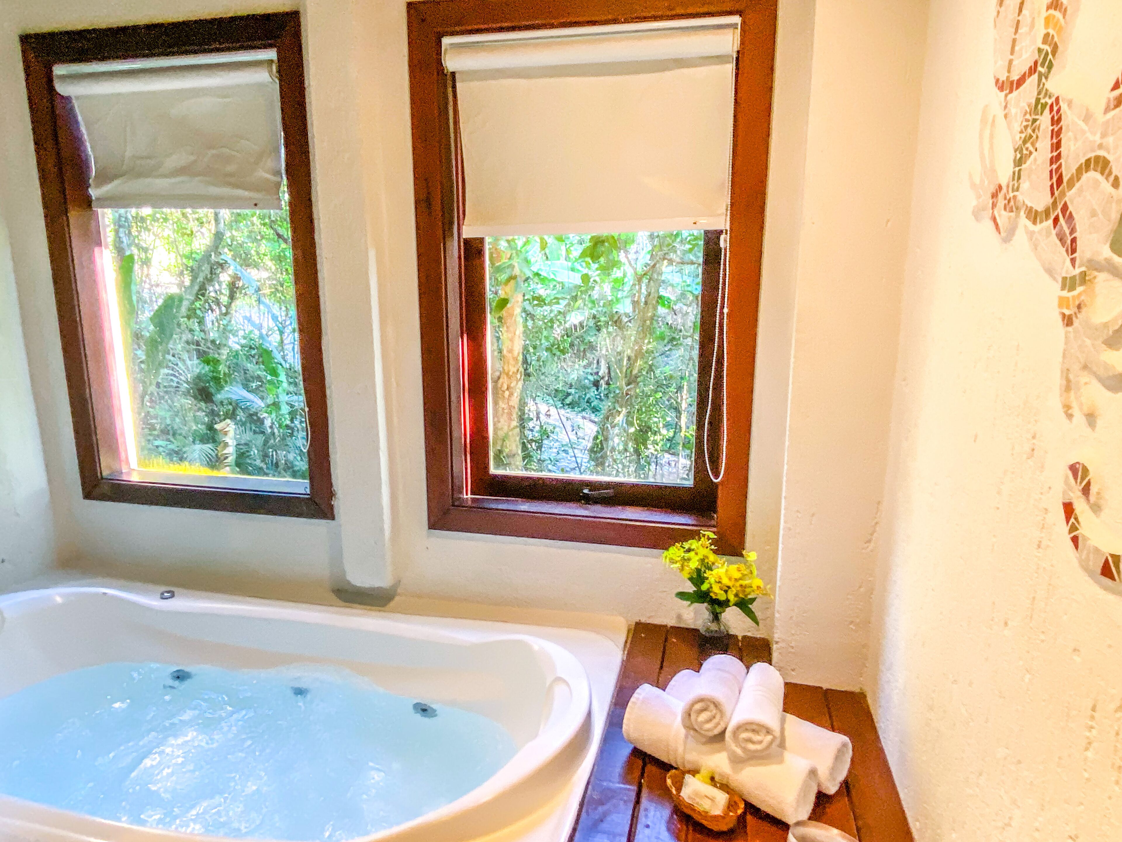 A bathroom with a bathtub, two windows with blinds, a wooden vanity, and a vase with yellow flowers.