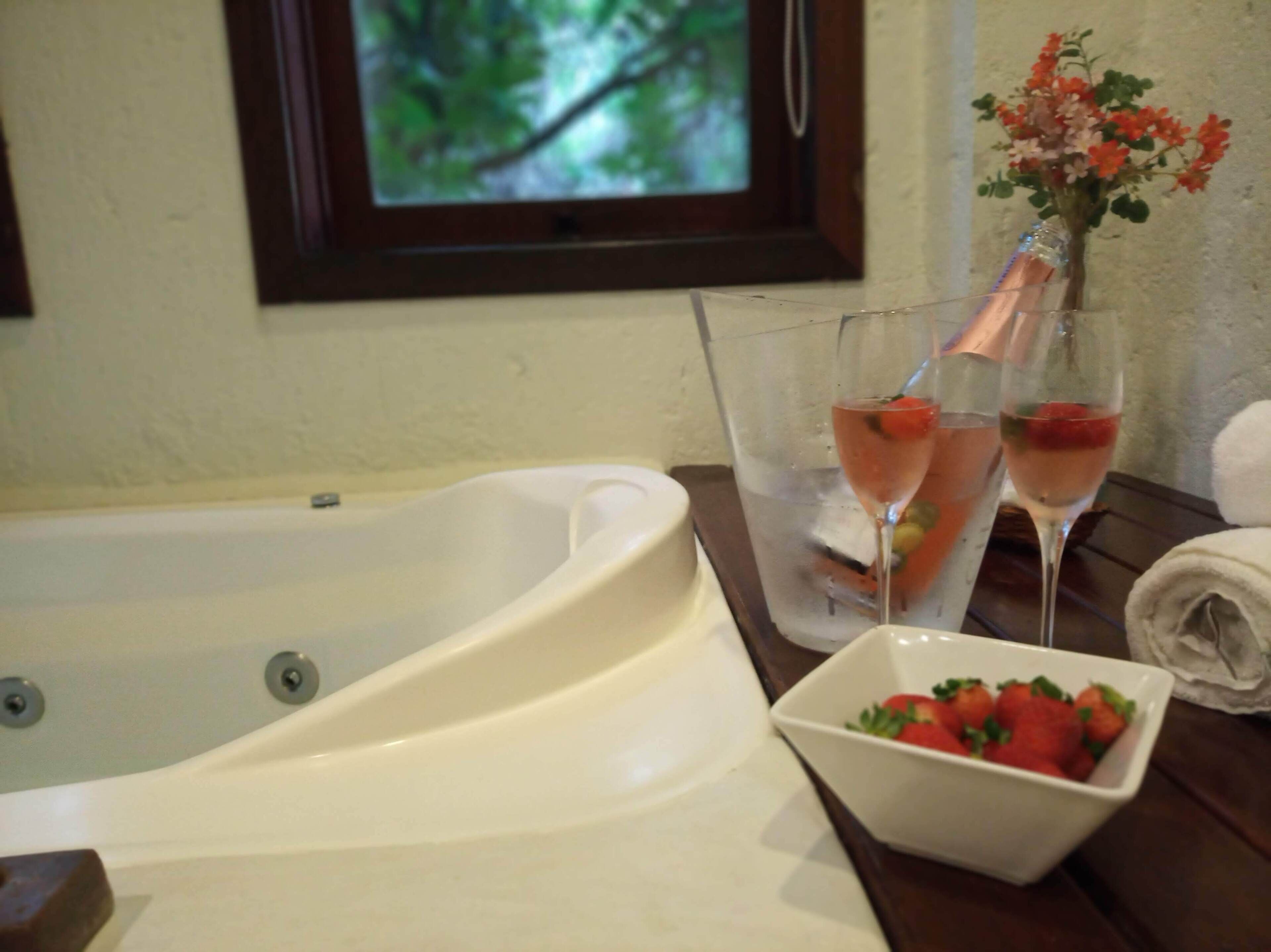 A bathroom with a bathtub, two glasses of pink drink, a bowl of strawberries, and a vase with flowers.