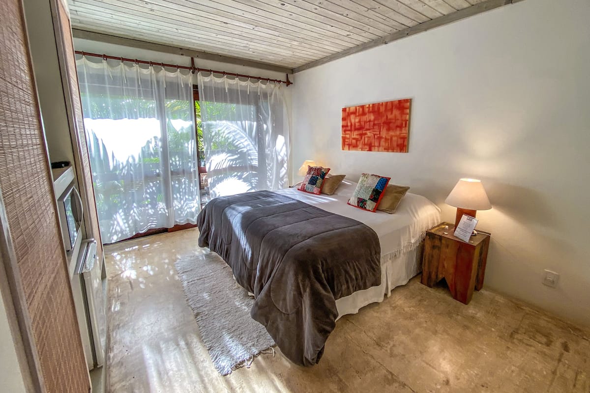 A bedroom with a bed, a nightstand, a lamp, a window with sheer curtains, and a painting on the wall.