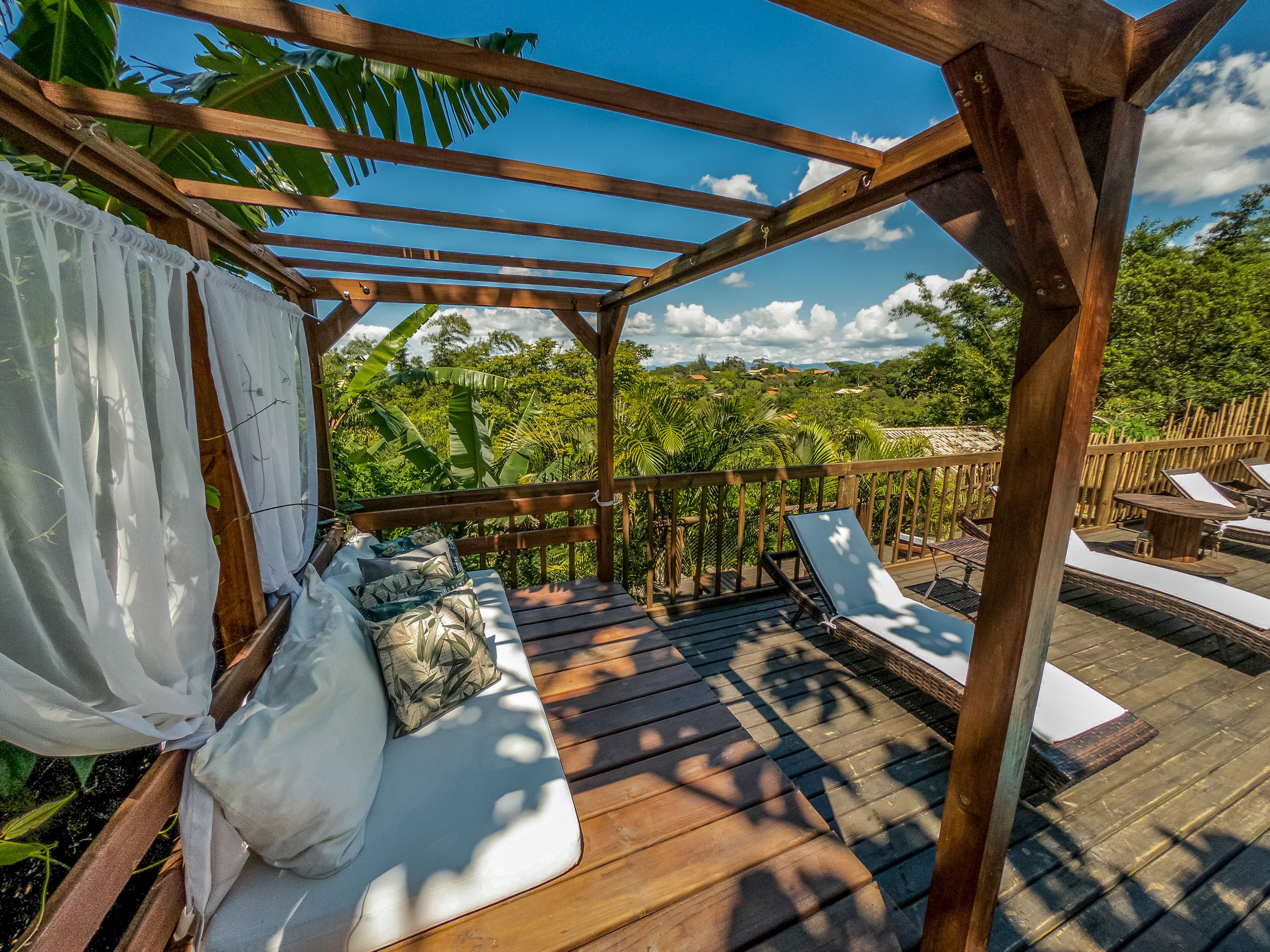 Una terraza de madera con zona de estar techada, cojines blancos y vistas a vegetación exuberante.