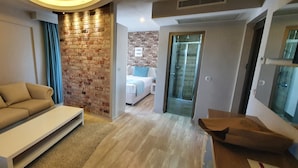 Een moderne woonkamer met een beige bank, een witte salontafel en een accentmuur van baksteen. Een glazen deur leidt naar een slaapkamer met een bed en een badkamer.