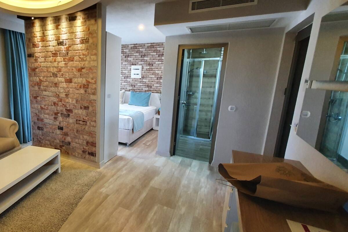 Un salon moderne avec un canapé beige, une table basse blanche et un mur en briques apparentes. Une porte vitrée donne accès à une chambre à coucher avec un lit et une salle de bain.