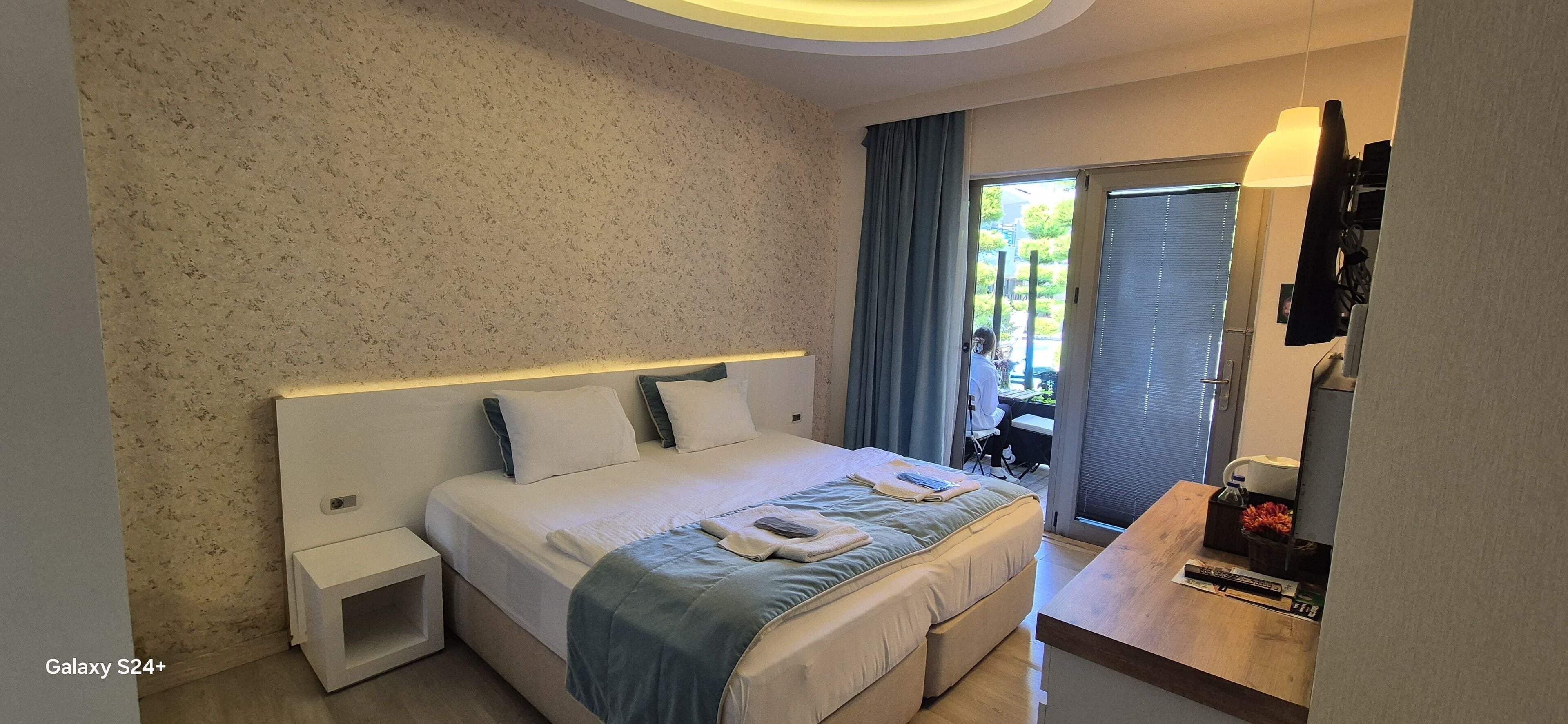 A modern hotel room with a large bed, bedside tables, a desk with a mirror, and a view of the outdoors.