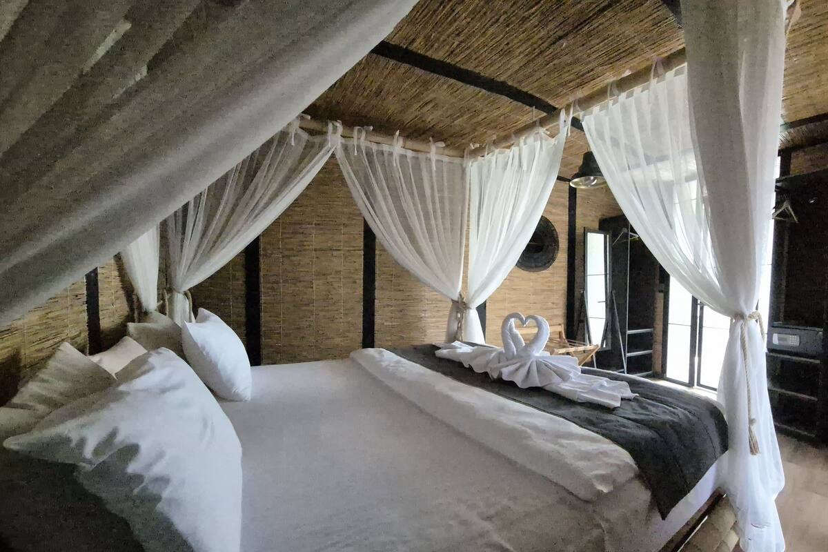 Un lit avec du linge de lit blanc et un baldaquin, une décoration en forme de cœur réalisée avec des serviettes, et un mur en briques rustiques.