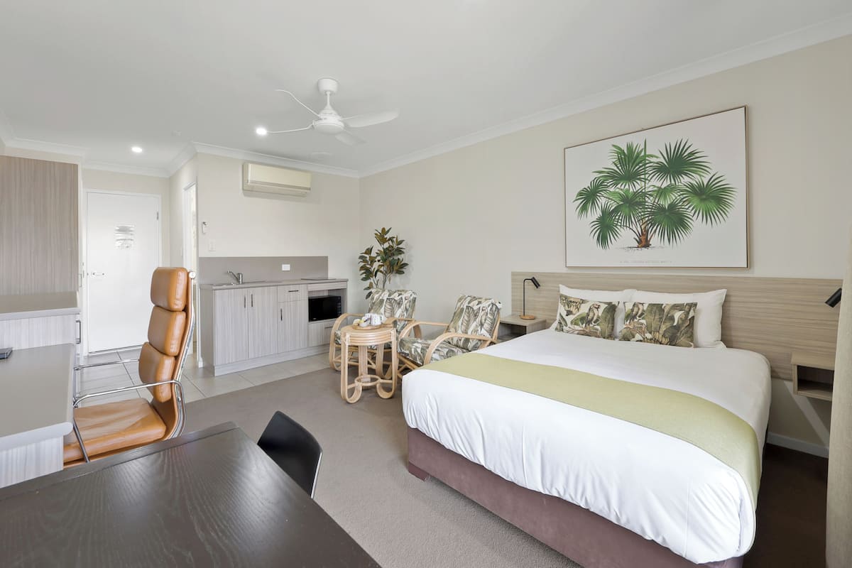 Quarto de hotel com cama, escrivaninha, cadeira, mesa de jantar, cozinha compacta e ventilador de teto.
