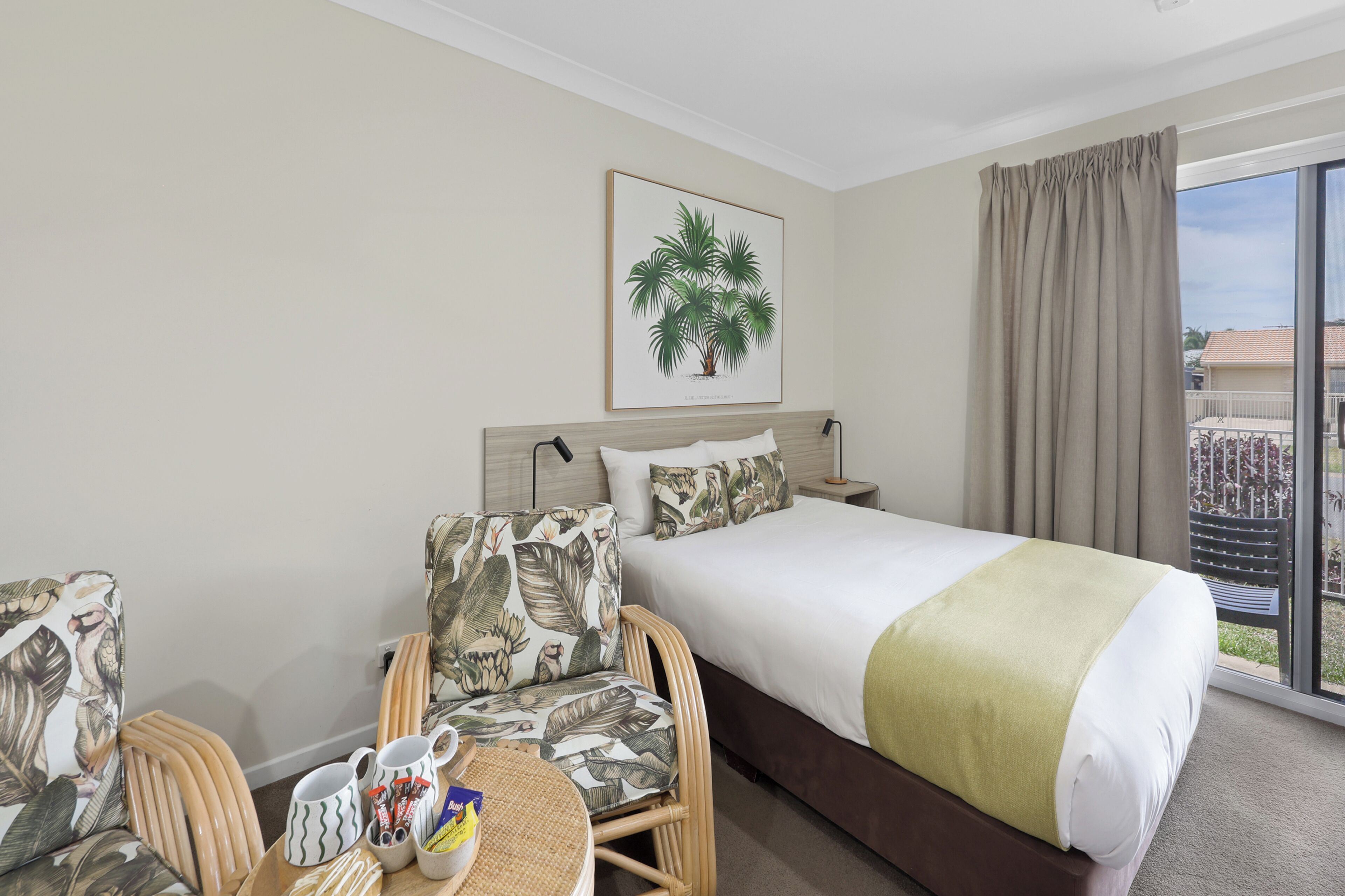 Une chambre d’hôtel avec un lit, deux chaises, une petite table avec un plateau, une plante en pot et une vue sur l’extérieur.