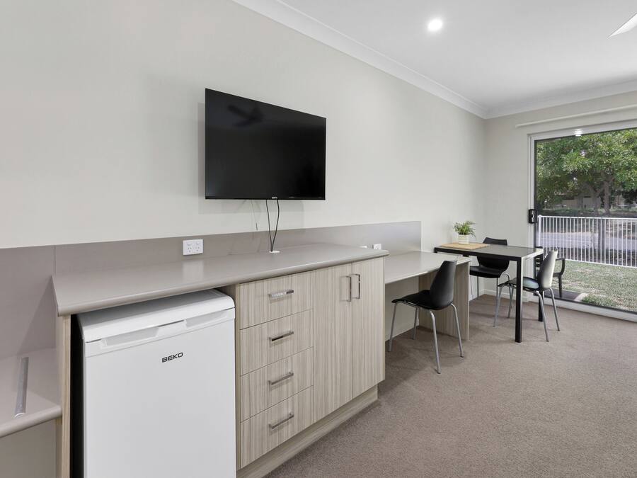 A kitchenette with a built-in dishwasher, a flat-screen TV mounted above the counter, a dining area with a table and chairs, and a sliding glass door leading to an outdoor area.