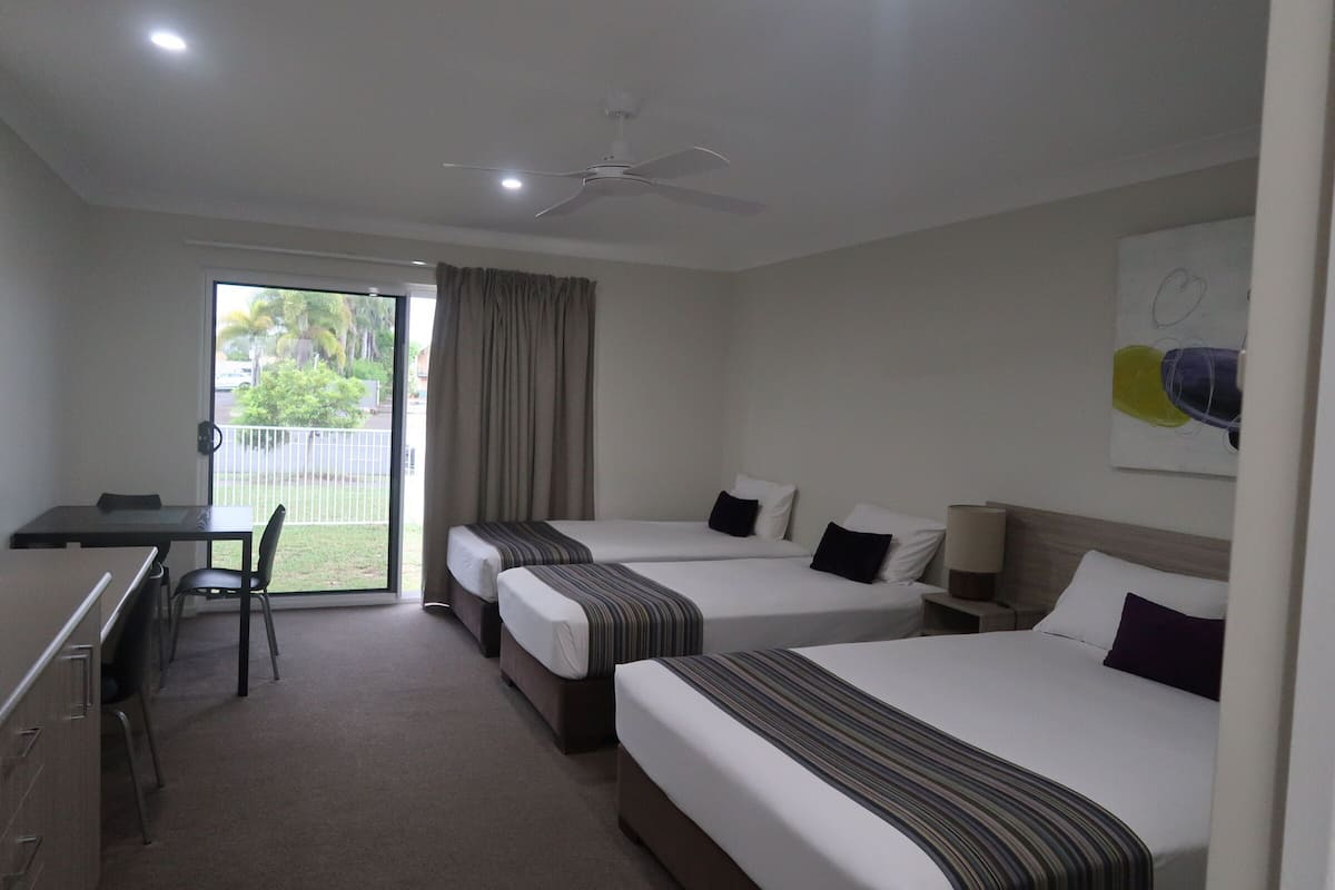 Quarto de hotel com duas camas de solteiro, uma escrivaninha e uma cadeira. Há um ventilador de teto e uma janela com cortinas.