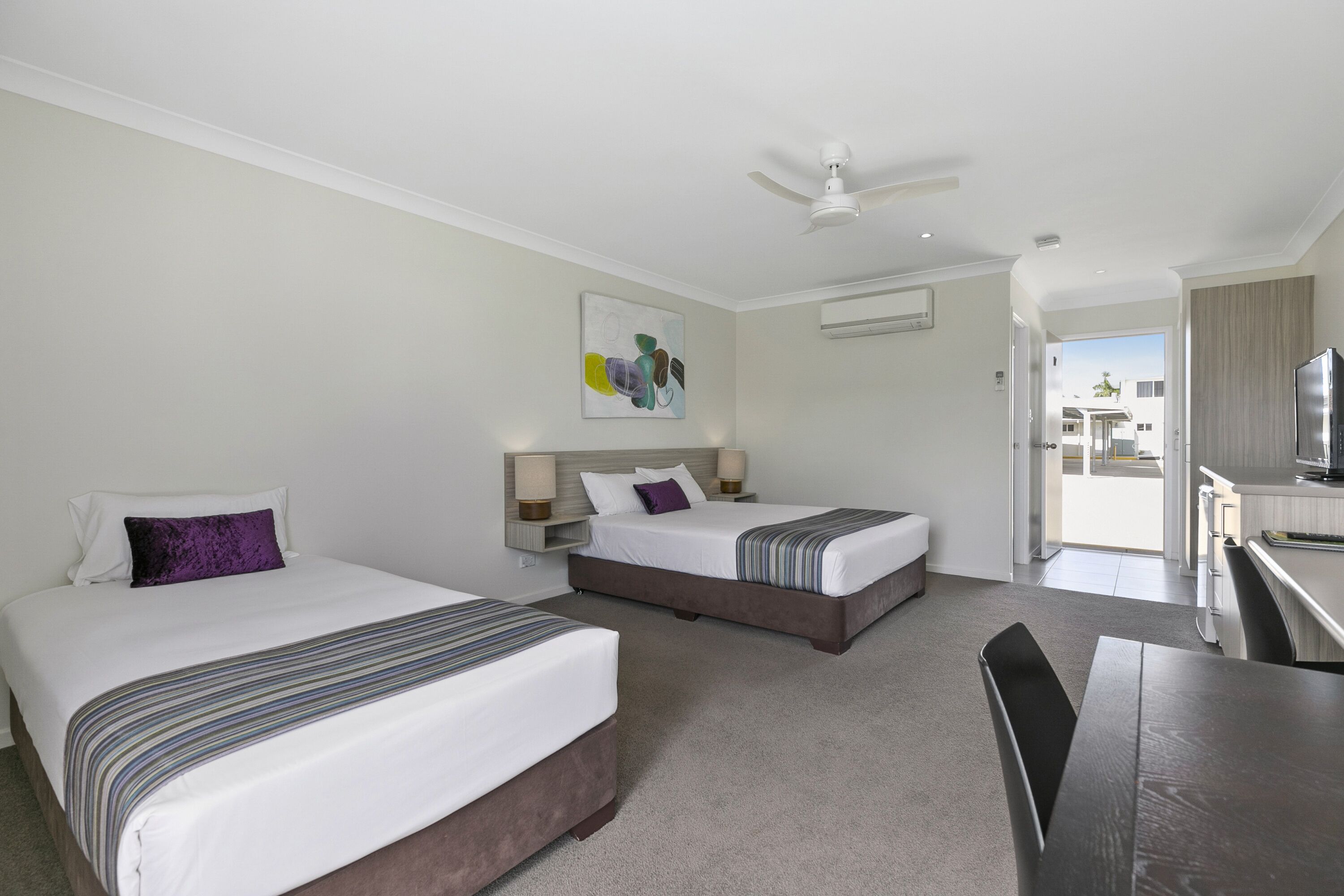 A hotel room with two beds, a ceiling fan, a painting on the wall, and a view of the outside.