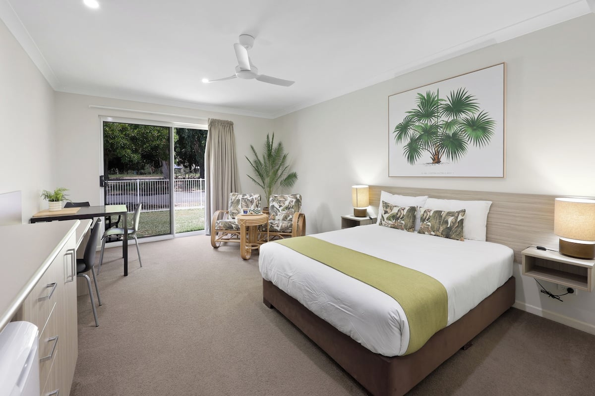 Quarto de hotel com cama, escrivaninha, cadeiras, ventoinha no teto, uma janela grande e uma planta.