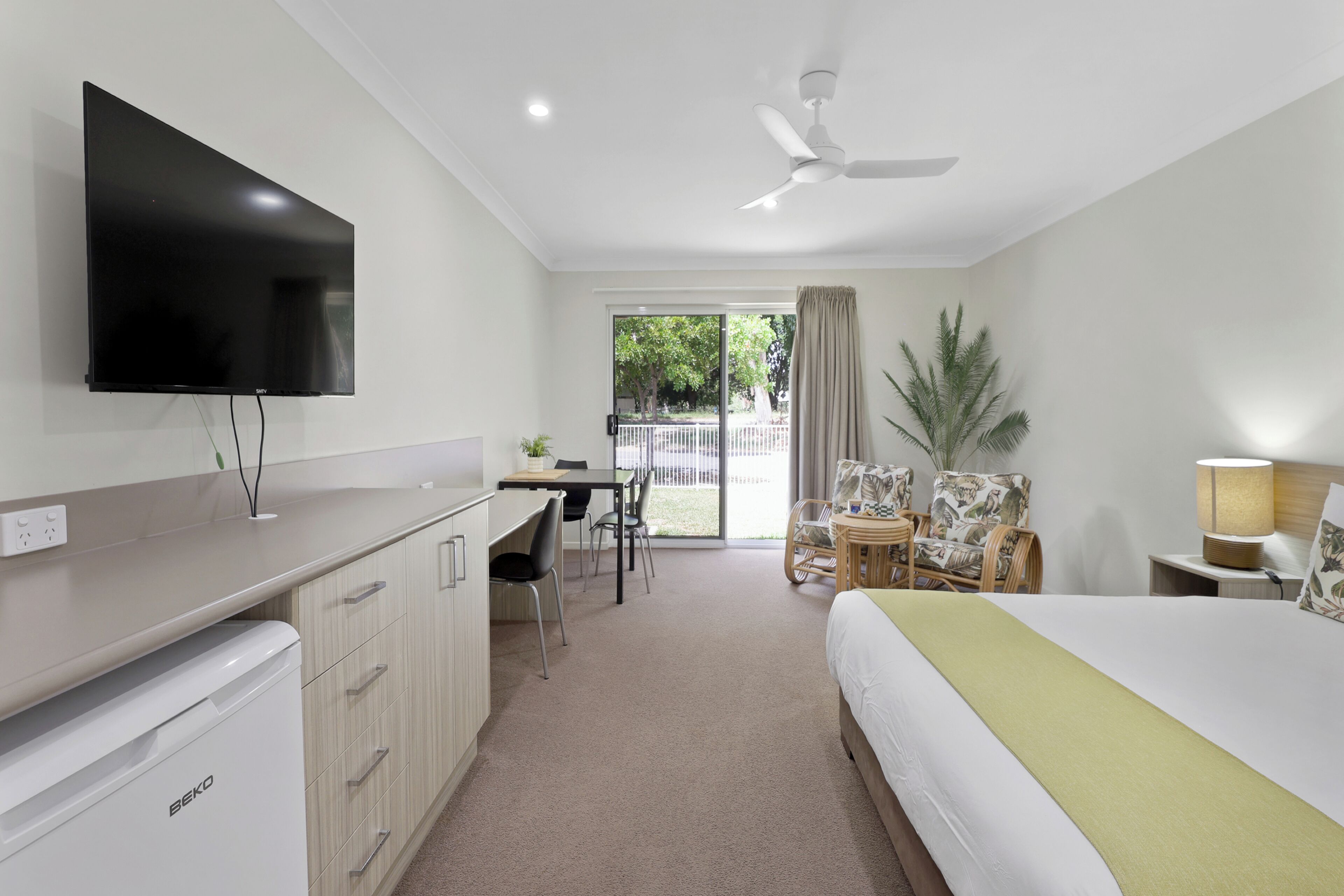 A hotel room with a bed, desk, chairs, a television, a ceiling fan, and a view of the outdoors.
