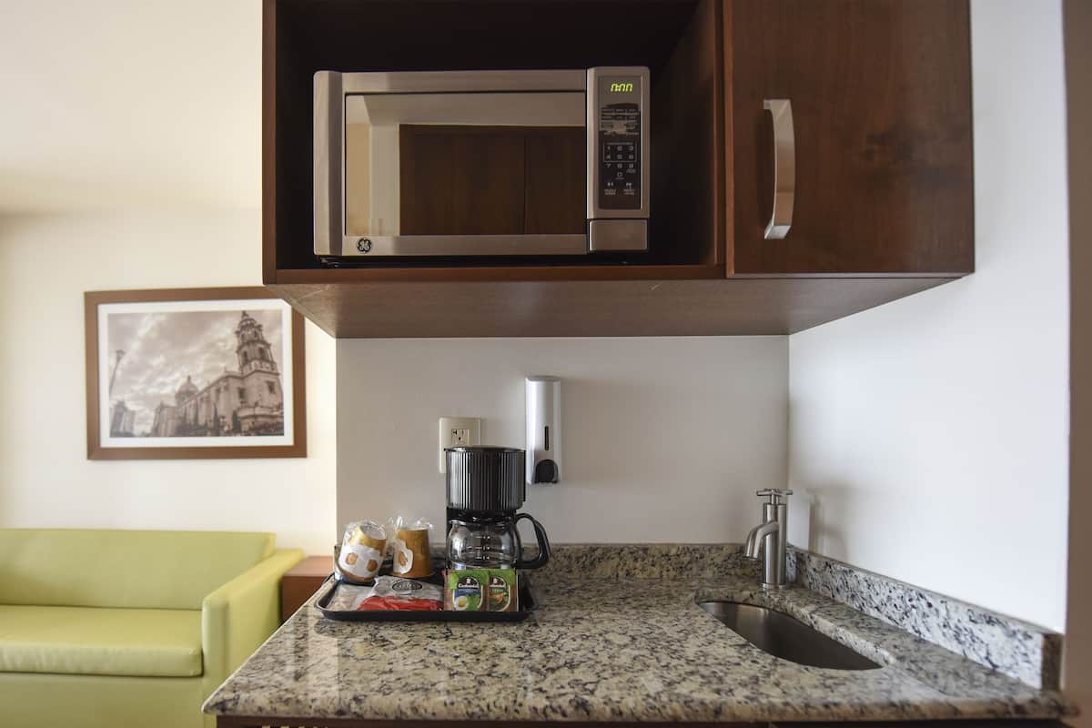 A compact kitchen with a microwave, coffee maker, and a small sink. There is a framed picture on the wall and a green chair in the room.