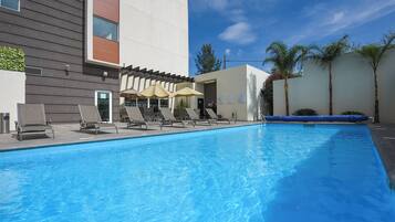 Una piscina con sillones de descanso y sombrillas, un edificio moderno con techo plano y palmeras.