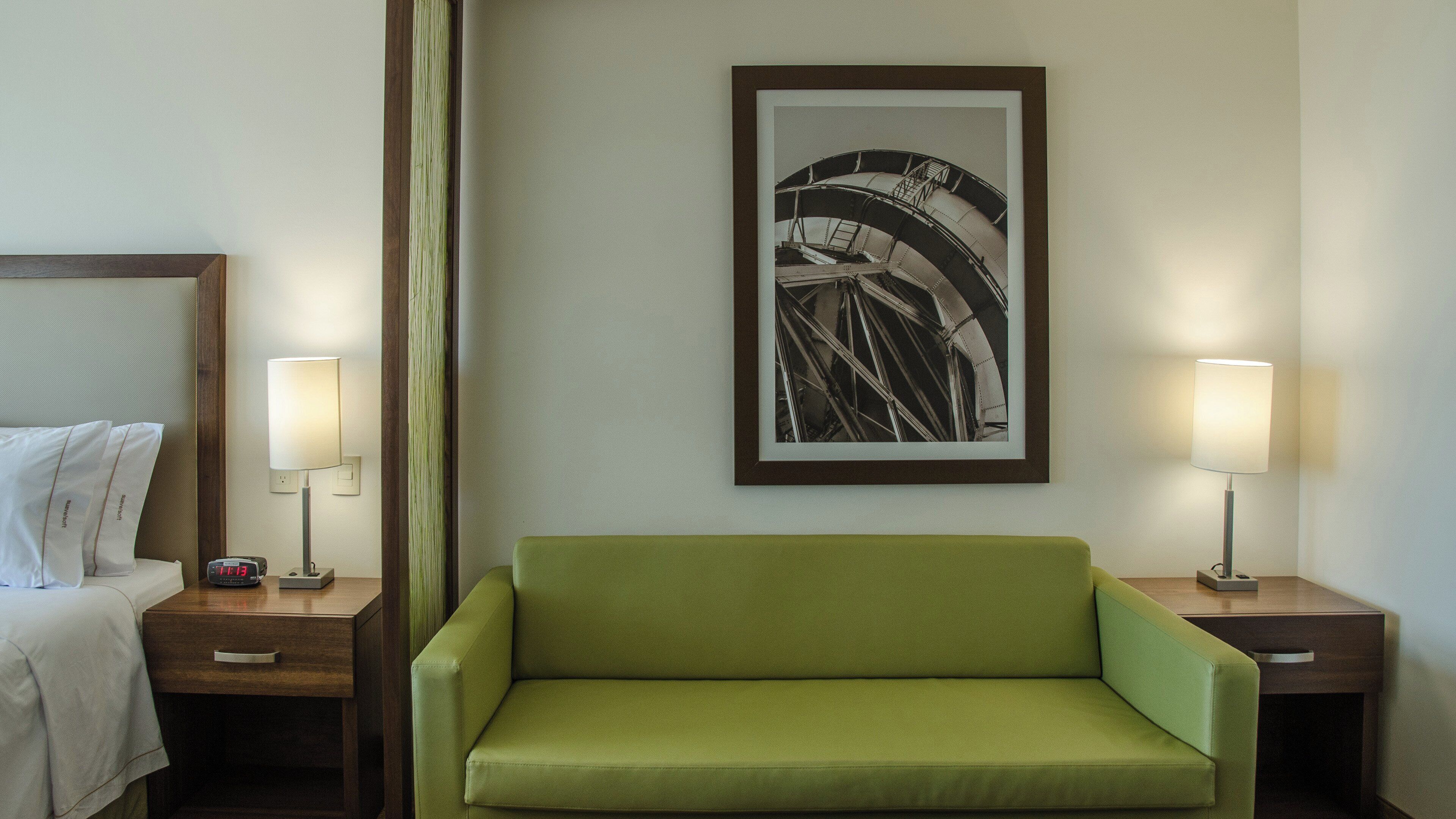 Habitación de hotel con cama, sofá verde, dos mesitas de noche, un cuadro en la pared y dos lámparas.