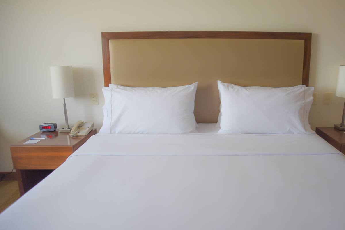 A hotel room with a large bed, two white pillows, a wooden headboard, two bedside tables with lamps, a bedside phone, and a remote control.