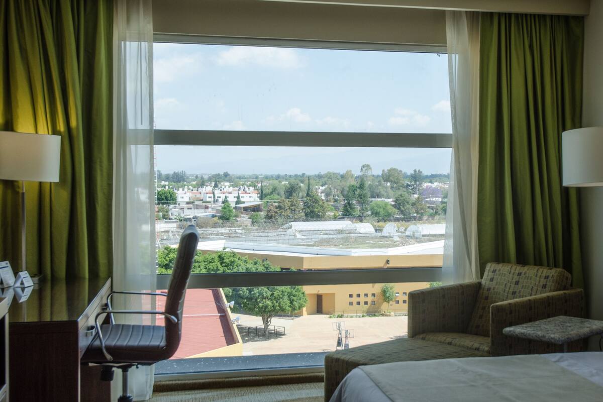 A hotel room with a bed, a chair, a desk, and a view of a cityscape through large windows.