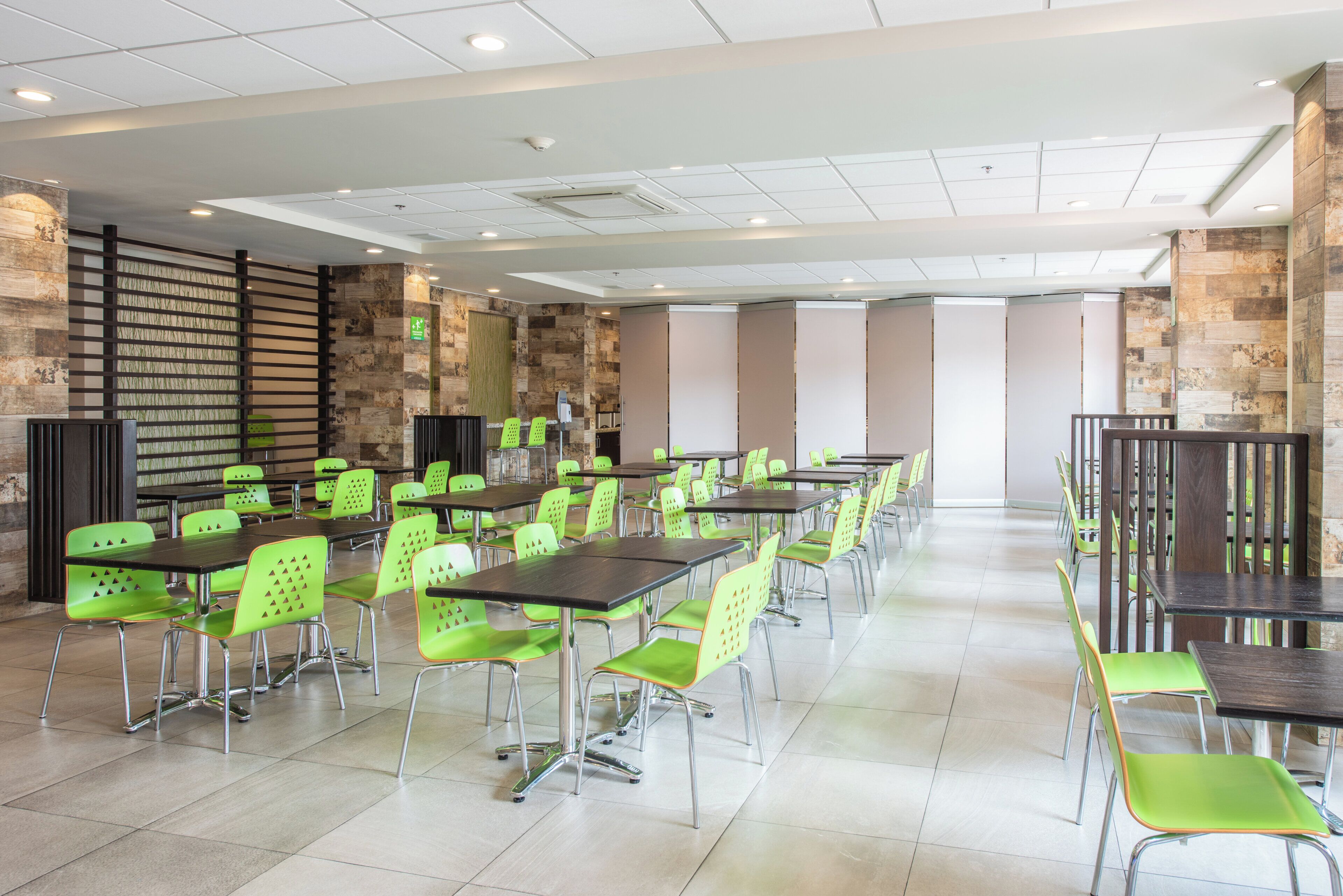 A spacious dining area with high tables and green chairs, a stone accent wall, and a ceiling with recessed lighting.