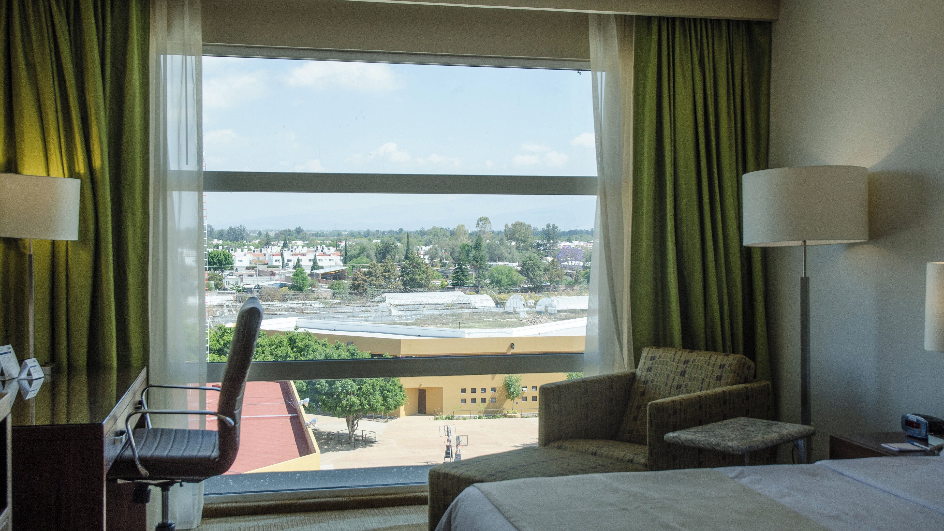 A hotel room with a bed, a chair, a desk, and a view of a cityscape through large windows.