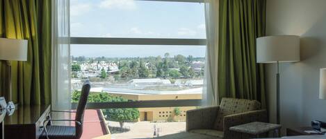 A hotel room with a bed, a chair, a desk, and a view of a cityscape through large windows.