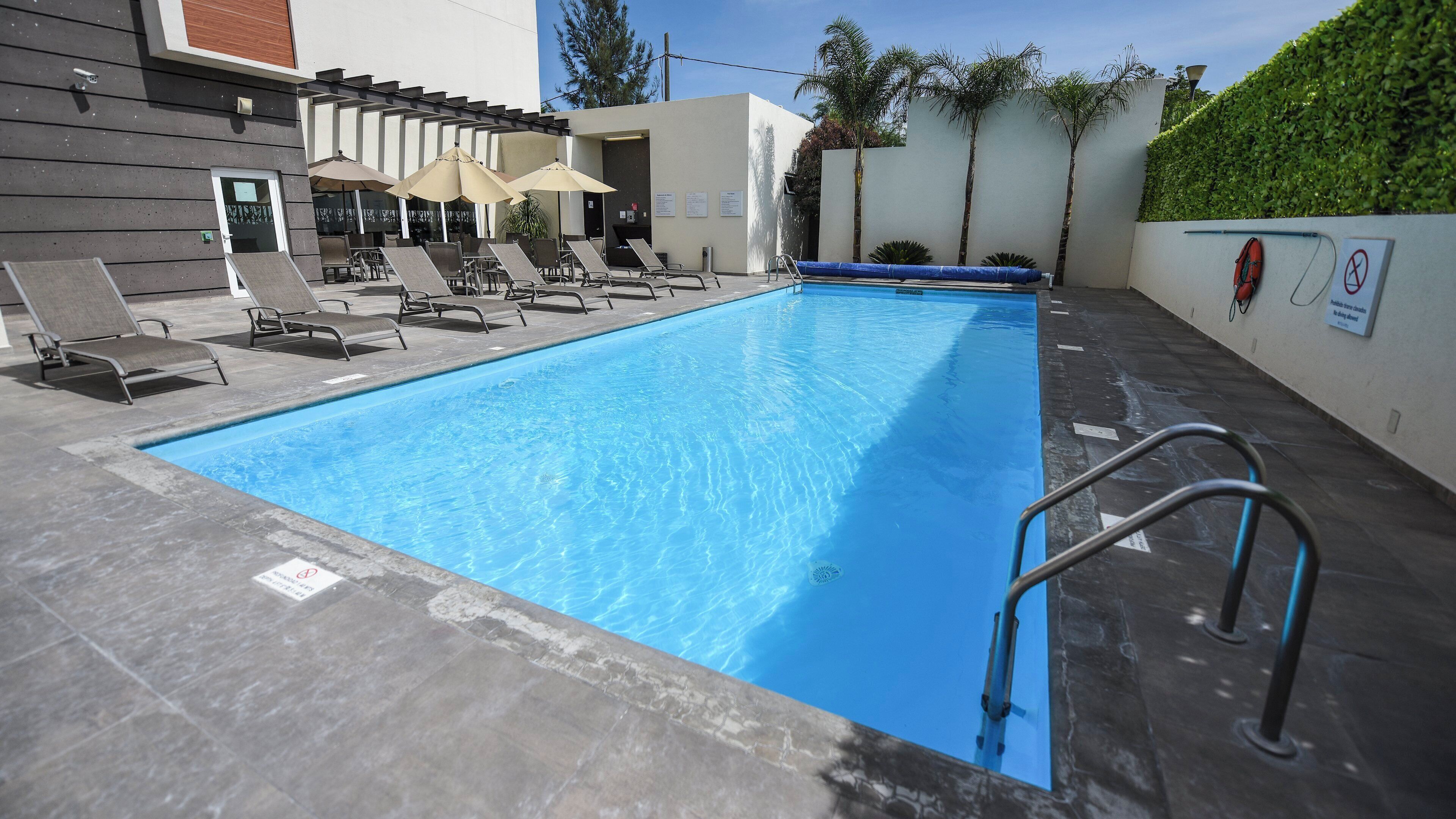 A swimming pool with lounge chairs and umbrellas, surrounded by a modern building with a tiled floor.